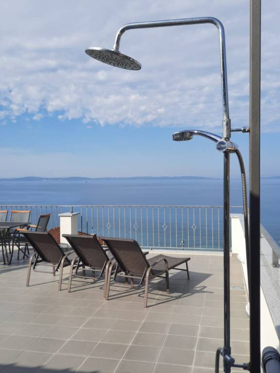 Terrasse mit Meerblick, ausgestattet mit Grill, Dusche, Liegestühlen, Tischen, Stühlen und Sonnenschirm.